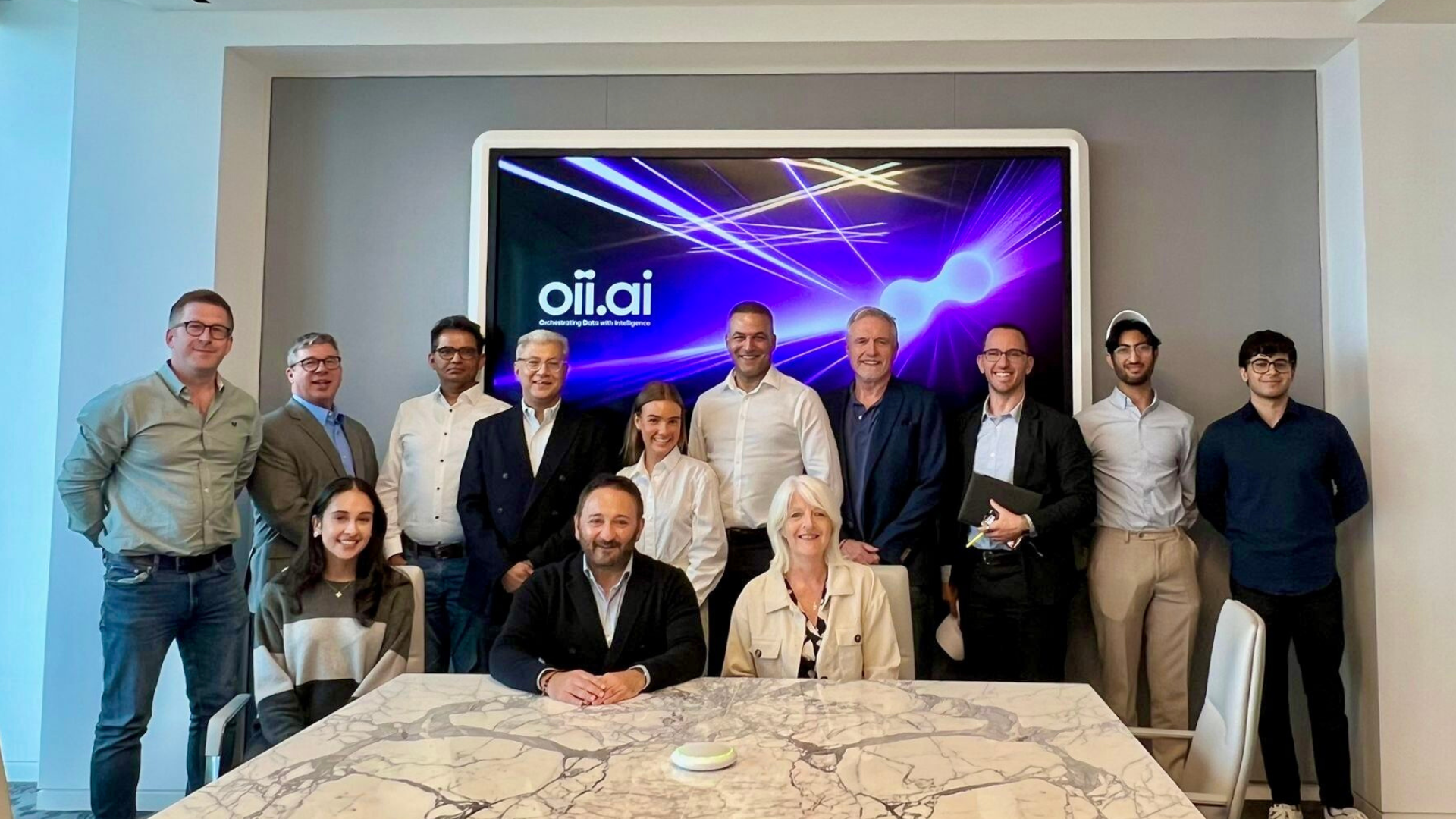 A group of 13 people, smiling in a modern conference room. A screen displaying "oii.ai" is behind them, creating a professional and positive tone.